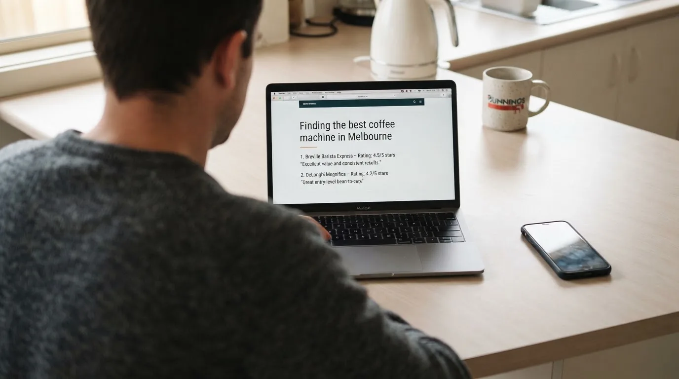 A person at a kitchen table scrolling through review pages on a laptop, phone face-down beside them.